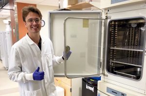 Julien wearing a white lab coat and blue gloves while opening the door of a lab incubator.