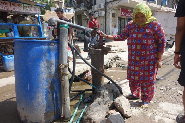 Photo of an urban street in India with a woman standing next to pipe coming out of the ground from which water is flowing into a blue barrel.