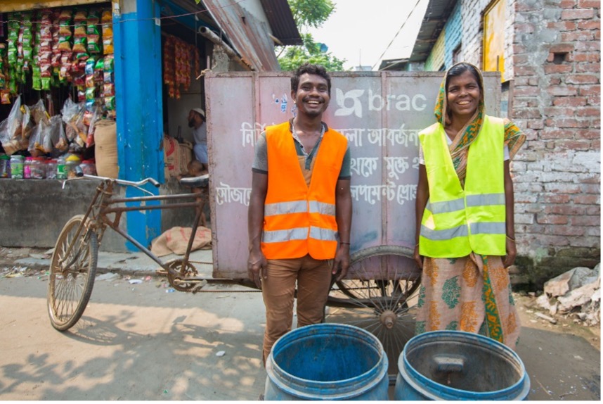 a male and female worker in Bangladesh smiling to camera while earing reflective safety vests and standing behind two plastic barrels on the side of a street, a transport bicycle and small store in the background