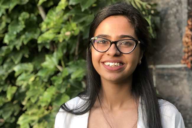 Khadija Rana looking to camera and smiling, standing in front of an ivy-covered wall