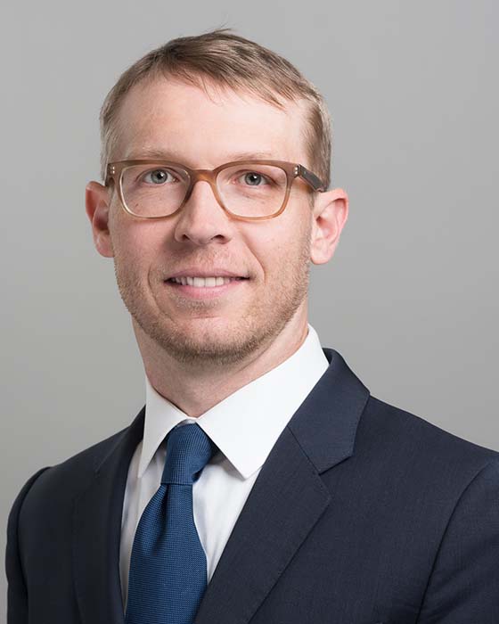 Professor Zeb Tate wearing light brown glasses and a dark blue suit looking to camera and smiling in front of a pale grey background