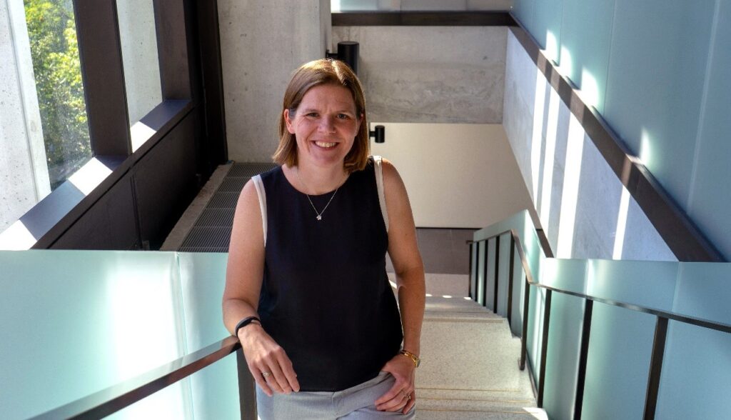 Prof Enright Jerger standing on a staircase with frosted glass and metal railings, looking up the stairs toward the camera and smiling