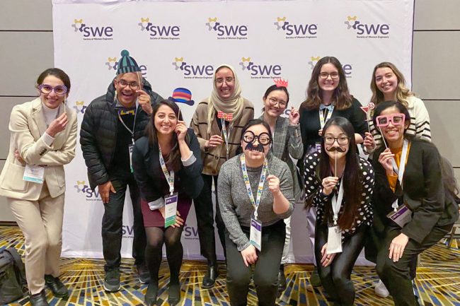 A group of mostly female students holding selfie props like glasses, hats and moustaches to their faces and laughing, while standing in front of a backdrop with the SWE logo on it.