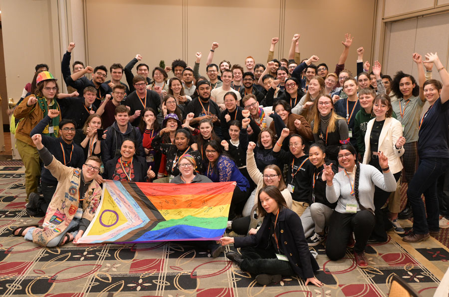 A large group of students, some sitting on the ground, some standing, holding a large Progress Pride flag.