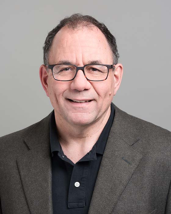 Professor Jonathan Rose wearing glasses, a black shirt and dark grey blazer, and smiling to camera in front of a pale grey background