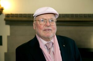 photo of K.C. Smith wearing a dark suit with shirt, tie, scarf, and cap in shades of pale pink
