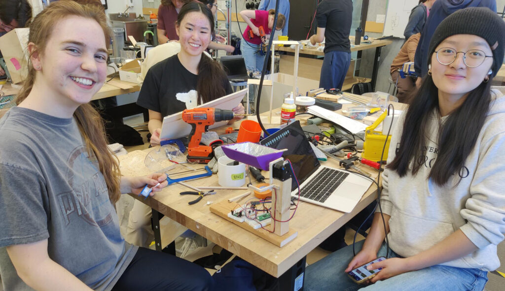 three female Praxis III students sitting at a wooden studio bench with an electric drill, various hand tools, a laptop, and their design prototype