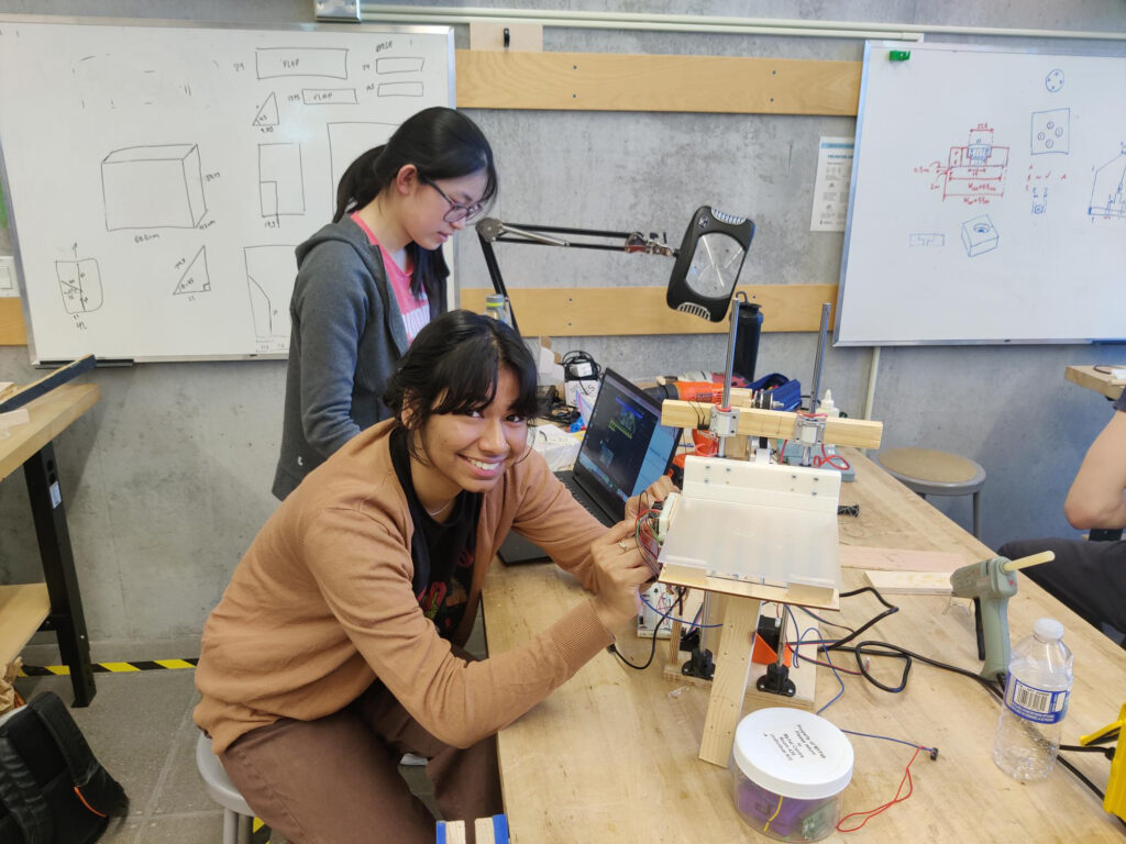 two female Praxis III students working on a prototype made of wood, metal, and plastic on a wooden bench in a design studio