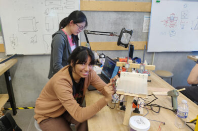 two female Praxis III students working on a prototype made of wood, metal, and plastic on a wooden bench in a design studio